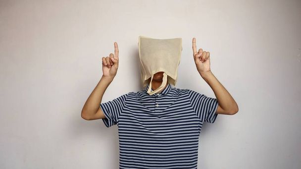 jeune homme asiatique portant un sac à provisions sur la tête avec un geste de la main pointant vers le haut avec un fond blanc isolé - Photo, image