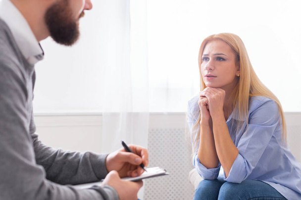 Молодая женщина сидит задумчиво, ее подбородок покоится на ее руке, вступает в разговор с мужчиной терапевтом, который делает заметки - Фото, изображение