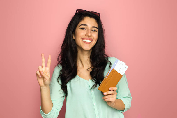 A cheerful young middle eastern woman, ready for travel, stands against a pink background holding her passport and boarding pass, flashing a peace sign with a beaming smile. - Photo, Image