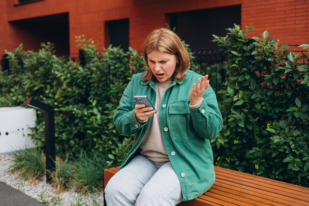 Retrato de mujer joven sosteniendo el celular en las manos en la calle en verano, mirando la pantalla con expresión irritada, cometió un error o se molestó con mensajes de texto y llamadas
 - Foto, Imagen