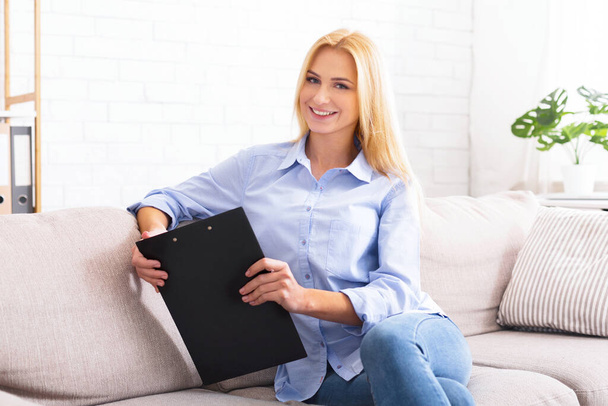 A woman is seated on a couch, holding a binder in her hands. She appears to be focused and engaged in the contents of the binder, possibly studying or working on a project. - Photo, Image