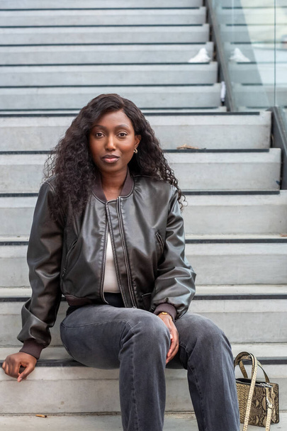 Een jonge zwarte vrouw zit terloops op betonnen treden in een stedelijke omgeving. Ze draagt een stijlvol zwart leren jasje over een witte tanktop, gecombineerd met vervaagde jeans, en een bruin en zwart geweven draagtas - Foto, afbeelding