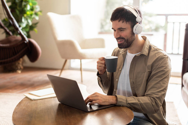 Een man zit aan een tafel, gefocust op zijn laptop scherm. Naast hem staat een kop koffie, stoom stijgt. De mensenhanden typen op het toetsenbord terwijl hij werkt.. - Foto, afbeelding