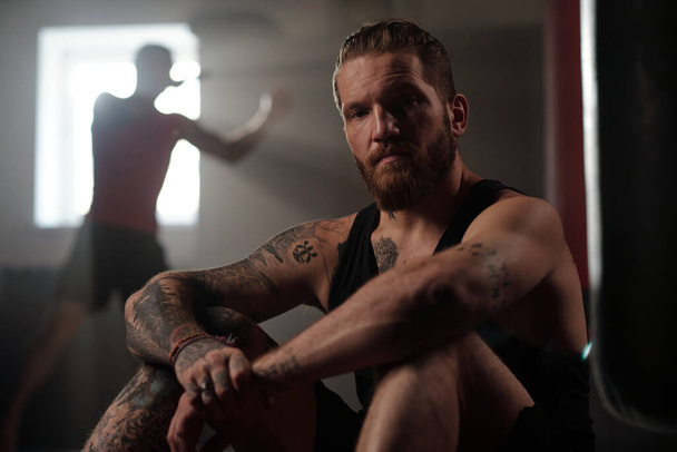 Βίαιος αθλητής με τατουάζ στα πόδια, τα χέρια, τους ώμους και το στήθος κάθεται στο πάτωμα του γυμναστηρίου κατά τύπος κλοτσιές σάκο του μποξ - Φωτογραφία, εικόνα