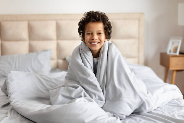 Ein afroamerikanisches Kind sitzt auf einem Bett, kuschelig in eine weiche Decke gewickelt. Die Kinderaugen sind geschlossen, was auf Schlaf oder Ruhe hindeutet. Der Raum ist schwach beleuchtet und schafft eine gemütliche Atmosphäre. - Foto, Bild