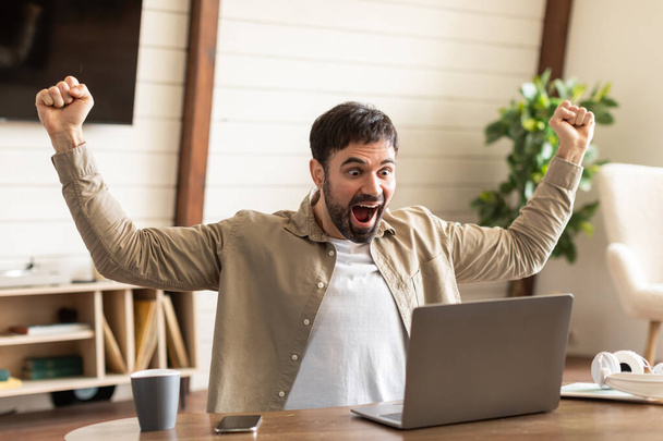 Ένας άντρας κάθεται μπροστά από το λάπτοπ του σε ένα γραφείο στο σπίτι, τα χέρια του σηκωμένα σε μια θριαμβευτική χειρονομία, φορώντας ένα απλό πουκάμισο. Το πρόσωπό του εκφράζει έντονο ενθουσιασμό και χαρά - Φωτογραφία, εικόνα
