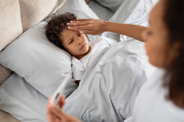 Znepokojená mladá afroamerická matka jemně položí ruku na své dětské čelo, aby zjistila, zda nemá horečku, zatímco dítě leží v posteli. Matka se zdá pozorná a starostlivá - Fotografie, Obrázek