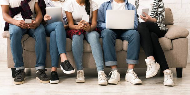 Обрізані п'ятьма багатоетнічними підлітками, які сидять поруч на зручному бежевому дивані, кожен з яких займається різними цифровими пристроями, включаючи смартфони, планшети та ноутбуки - Фото, зображення