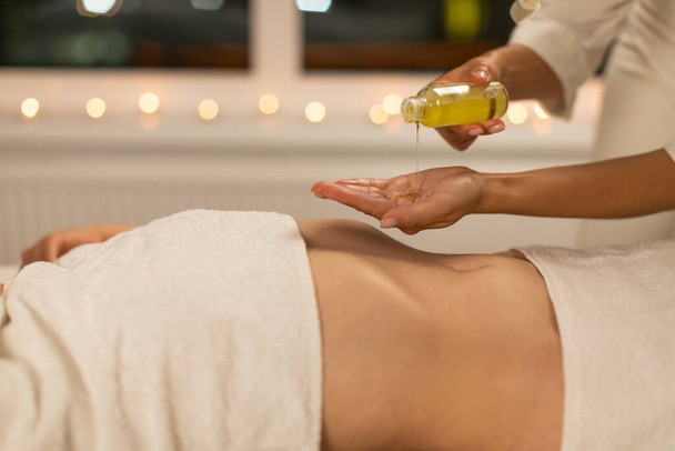In a serene spa setting with ambient lighting, a professional masseuse is captured preparing for a soothing massage session by pouring massage oil onto their hands while the client waits - Photo, Image