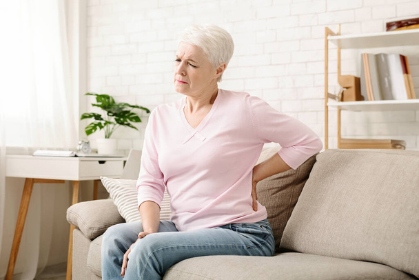 Mujer mayor que sufre de dolor de espalda, sentado en el sofá y tocando el lomo en casa, espacio vacío
 - Foto, Imagen
