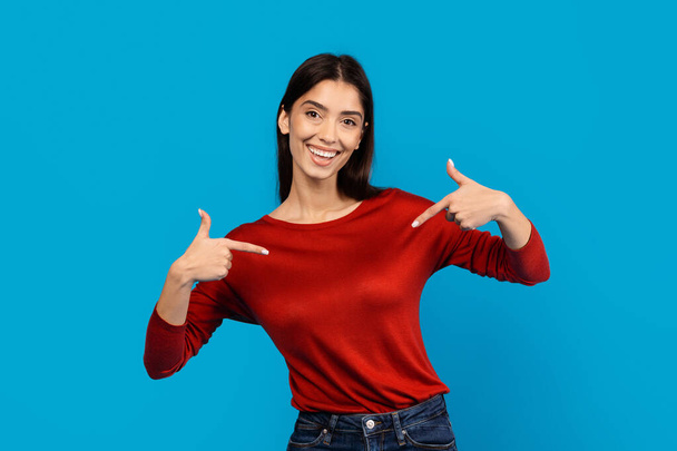 Egy mosolygó fiatal nő, alkalmi piros, hosszú ujjú felsőben, egy szilárd kék háttér előtt áll. A magabiztosság kifejezésével, mindkét hüvelykujját önmaga felé fordítja. - Fotó, kép