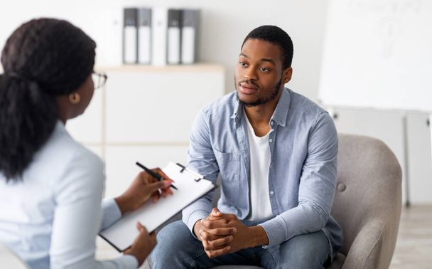 Boldogtalan, fiatal fekete férfi, aki pszichológussal találkozik egy elmegyógyintézetben. Pszichoterapeuta jegyzetel beszélgetés közben depressziós férfi beteg - Fotó, kép