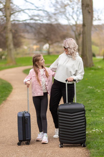 Woman and girl smiling and posing with suitcases against the backdrop of a spring park - Photo, Image