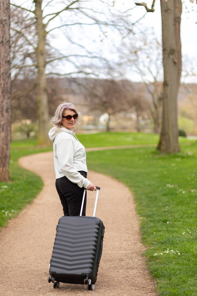 Mujer con una maleta sonríe mirando por encima del hombro en un callejón del parque - Foto, imagen