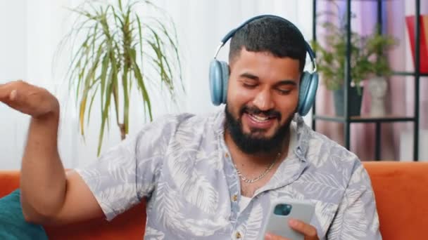 Щасливий розслаблений індійський арабський чоловік в бездротових навушниках танцює на дивані вдома, вибираючи слухати улюблену енергійну диско поп-музику в смартфоні. Діяльність вихідних людей - Кадри, відео