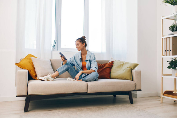 Mujer sonriente sosteniendo el teléfono móvil en la acogedora sala de estar, disfrutando de juegos en línea - Foto, imagen