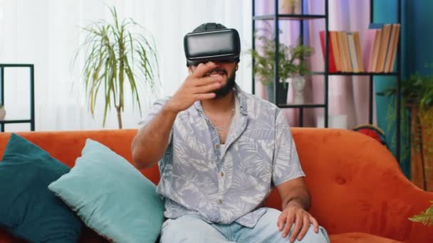 Indian man using virtual reality futuristic technology VR app headset helmet to play simulation 3D 360 online video game, watching film movie at modern home apartment. Guy in goggles sitting on sofa - Footage, Video