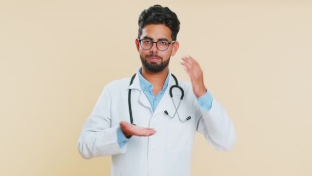 Potrzebuję więcej, proszę, daj mi. Indyjski młody lekarz kardiolog wykazujący się odrobiną gestu, mierzący małe rozmiary, błagający o pomoc. Arabian aptekarz facet izolowany na beżowym tle - Materiał filmowy, wideo