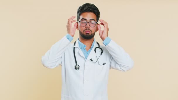 Wyczerpany zmęczony indyjski młody lekarz kardiolog człowiek zdejmuje okulary, czuje ból oczu, będąc przepracowane wypalenie od długich godzin pracy. senny wyczerpany facet tarcie oczy odizolowane na beżowy tło - Materiał filmowy, wideo