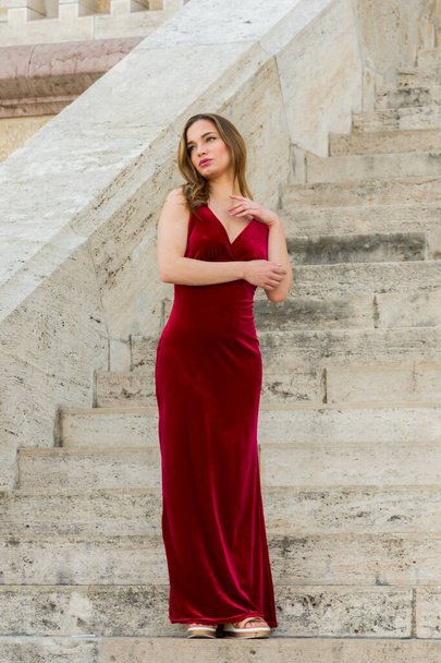A beautiful woman wearing red satin evening gown stands on the steps of an old building with her arms across her body - Photo, Image