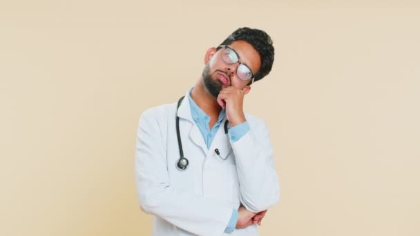 Sad Indian young doctor cardiologist man tired bored exhausted indifferent expression, not interested in communication talk, displeasure. Arabian apothecary pharmacy guy isolated on beige background - Footage, Video