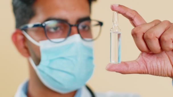 Indischer Arzt Kardiologe Mann in einer medizinischen Schutzmaske hält eine Ampulle Impfstoff Medikament Behandlung Injektion. Labortests. Arabische Apotheke Apotheker Kerl isoliert auf beigem Hintergrund - Filmmaterial, Video