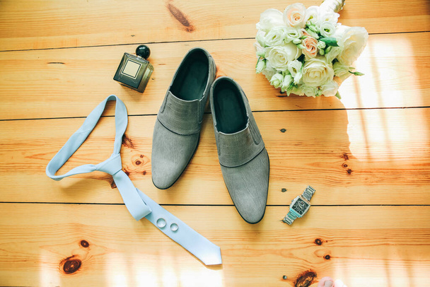 A vőlegény szürke cipője, kék nyakkendő és óra a fa háttéren. Esküvői részletek fotó. Csokor, gyűrű és parfümtartozék. - Fotó, kép