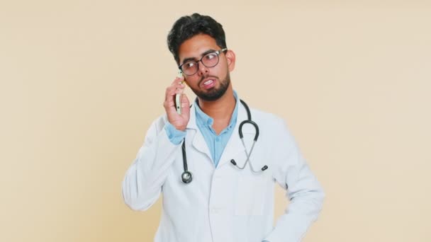 Zdenerwowany, zestresowany, zmęczony indyjski lekarz kardiolog, krzyczący przez dwa telefony komórkowe, kłóci się. Arabian aptekarz facet izolowany na beżowym tle - Materiał filmowy, wideo