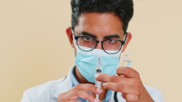 Indian young doctor cardiologist man holds syringe needle and ampoule tube with medical vaccine medicine treatment injection ready to use. Immunization. Arab scientist guy isolated on beige background - Footage, Video