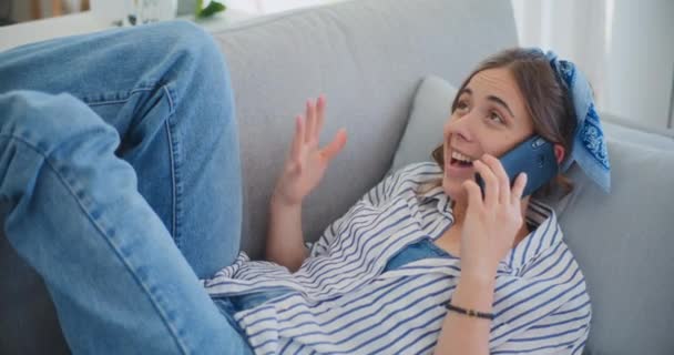 Een vrolijke vrouw ligt comfortabel op de bank in de woonkamer, kletst vrolijk op haar mobiele telefoon, straalt warmte en geluk uit tijdens het gesprek - Video
