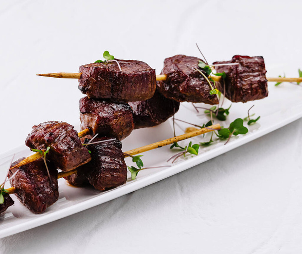 Sappige biefstuk blokjes op spiesjes versierd met verse kruiden, geserveerd op elegante witte schotel - Foto, afbeelding