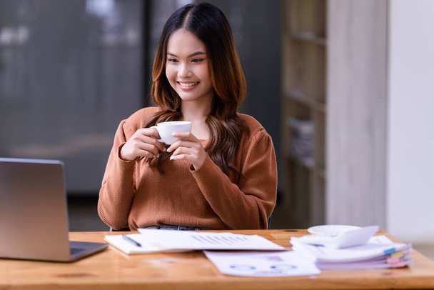 Nuori nainen, joka pitelee kuppia kahvia ja rentoutuu toimistossa työpöydän ääressä. Liiketoiminta rento käsite - Valokuva, kuva