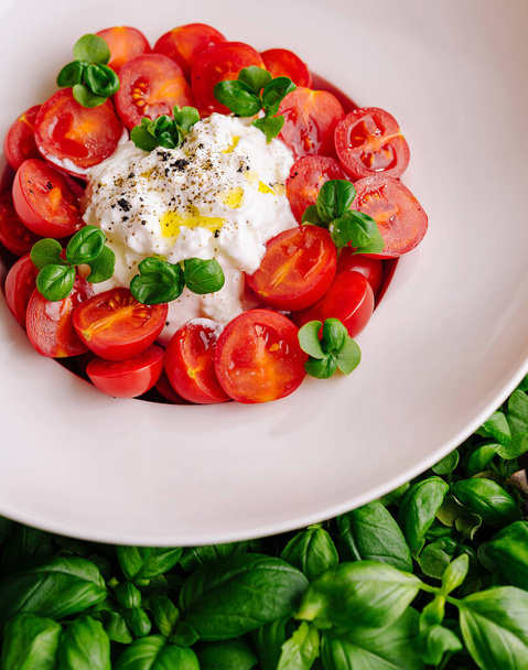 Dilimlenmiş kiraz domatesleri, kremalı burrata ve beyaz tabakta taze fesleğen yaprakları. - Fotoğraf, Görsel