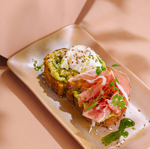 Elegante avocado toast belegd met gezouten prosciutto, een gepocheerd ei en verse kruiden op een stijlvol bord - Foto, afbeelding
