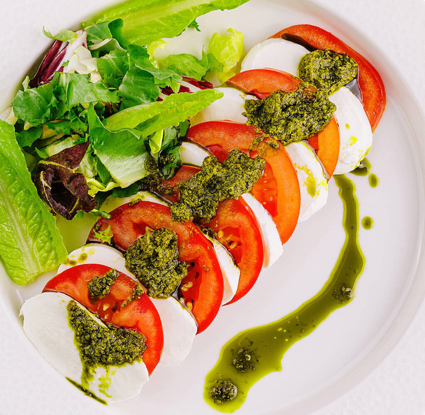Vue aérienne de la salade de caprese avec tomates mûres, mozzarella, pesto au basilic et légumes verts sur assiette blanche - Photo, image