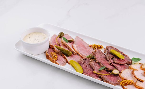 Gourmet platter of sliced deli meats served with pickles and a creamy dip on a white background - Photo, Image
