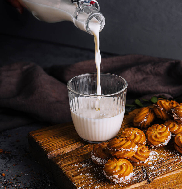 Flux de lait d'un pichet remplissant un verre, avec de délicieux biscuits sur une planche de bois - Photo, image