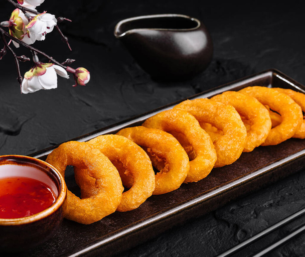 Золотисто-коричневые луковые кольца подаются на подносе с пряным соусом и палочками для еды, элегантная презентация продуктов питания - Фото, изображение