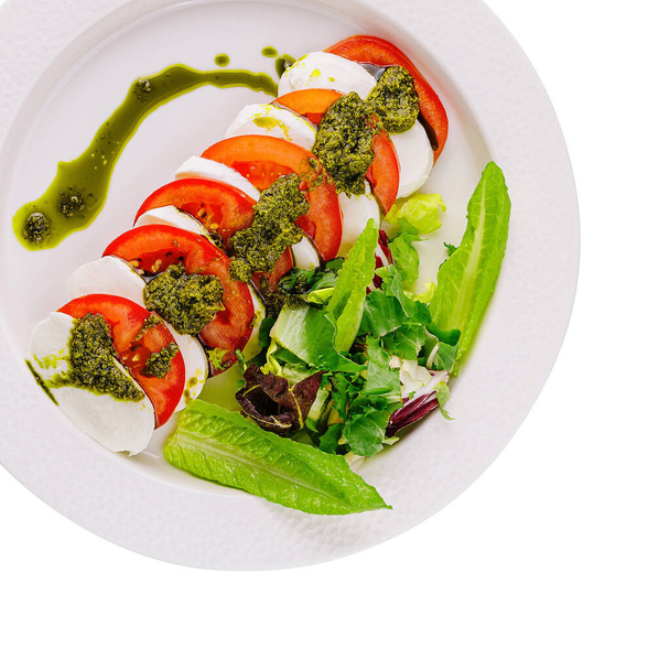 Olgun domatesli caprese salatası, mozzarella, fesleğen sosu ve beyaz tabakta yeşillikler. - Fotoğraf, Görsel