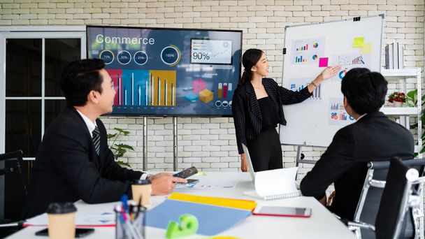 A cheerful and confident Asian businesswoman stands, present bar charts data from projector screen to her office colleagues. Asian business women leader role at the meeting. - 写真・画像