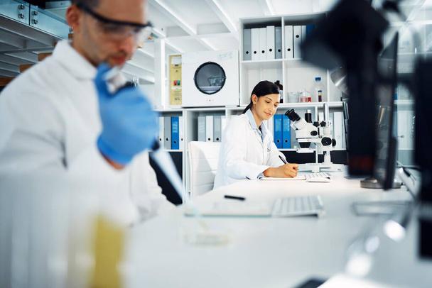 Scientist, woman and writing notes for research, test and chemical analysis for health innovation in lab on microscope. Dropper, science and medical study of drugs, liquid and exam for biotechnology. - Photo, Image