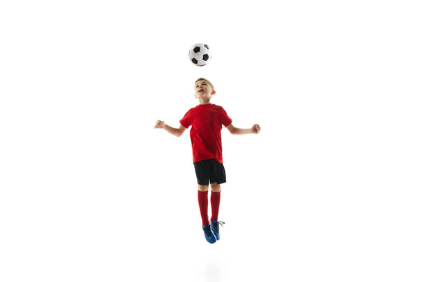 Imagen dinámica del niño en el aire, vestido con camiseta de fútbol rojo y pantalones cortos negros, dirigiendo hábilmente la pelota de fútbol sobre fondo blanco. Concepto de deporte profesional, campeonato, liga juvenil. Anuncio - Foto, imagen