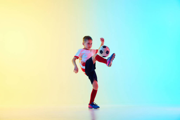 Dynamic portrait of little sporty boy with soccer ball kicking ball with knee in motion in neon light against gradient background. Concept of professional sport, championship, youth league, hobby. Ad - Photo, Image