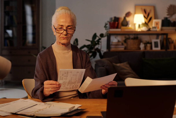 Femme âgée en lunettes prenant la facture financière pliée hors de l'enveloppe avant de la lire tout en étant assis à table devant un ordinateur portable - Photo, image