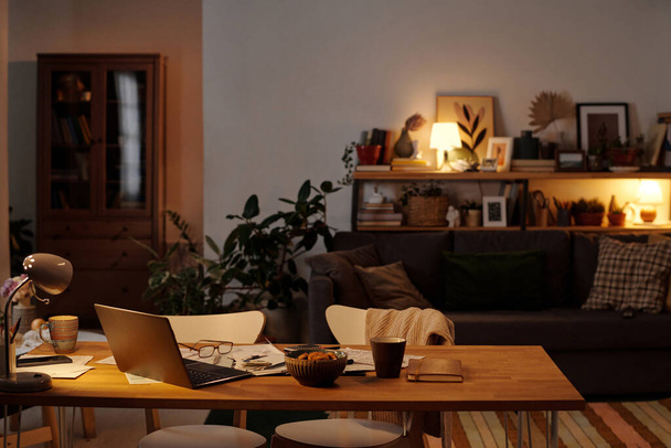 Parte de la amplia sala de estar con mesa de madera con computadora portátil, tazón con galletas caseras, taza de té o café y facturas financieras - Foto, Imagen