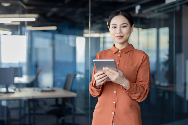 A professional Asian businesswoman looks directly at the camera while holding a digital tablet in a well-lit, modern office environment, symbolizing tech-savvy business operations. - Zdjęcie, obraz