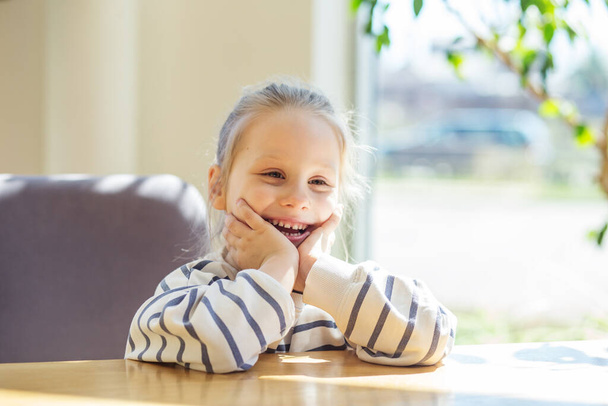Potěšená holčička sedící u dřevěného stolu, usmívající se s rukama na tvářích, ozářená přirozeným slunečním světlem. - Fotografie, Obrázek