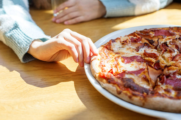 Person's hand reaching to grab slice of meat lover's pizza with crispy bacon and melted cheese on sunny table. - Photo, Image