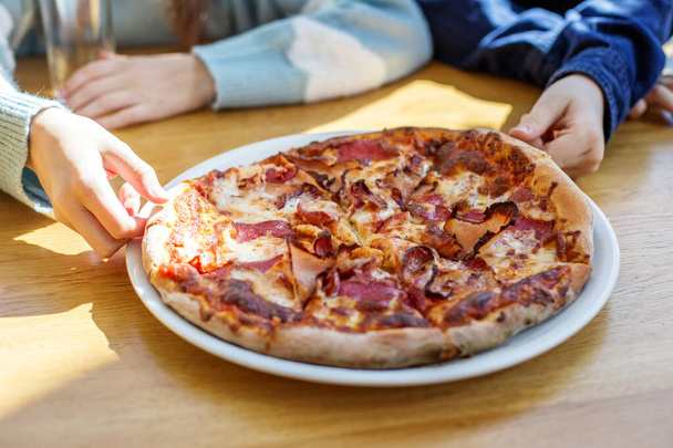 Family hands reaching to take slices of delicious meat lover's pizza on sunny wooden table, sharing meal together. - Photo, Image