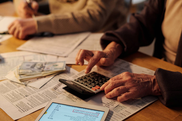 Зморшкуваті руки старшої невпізнаваної жінки, що тримає переслідувача над кнопкою калькулятора під час перевірки сум у фінансових рахунках - Фото, зображення